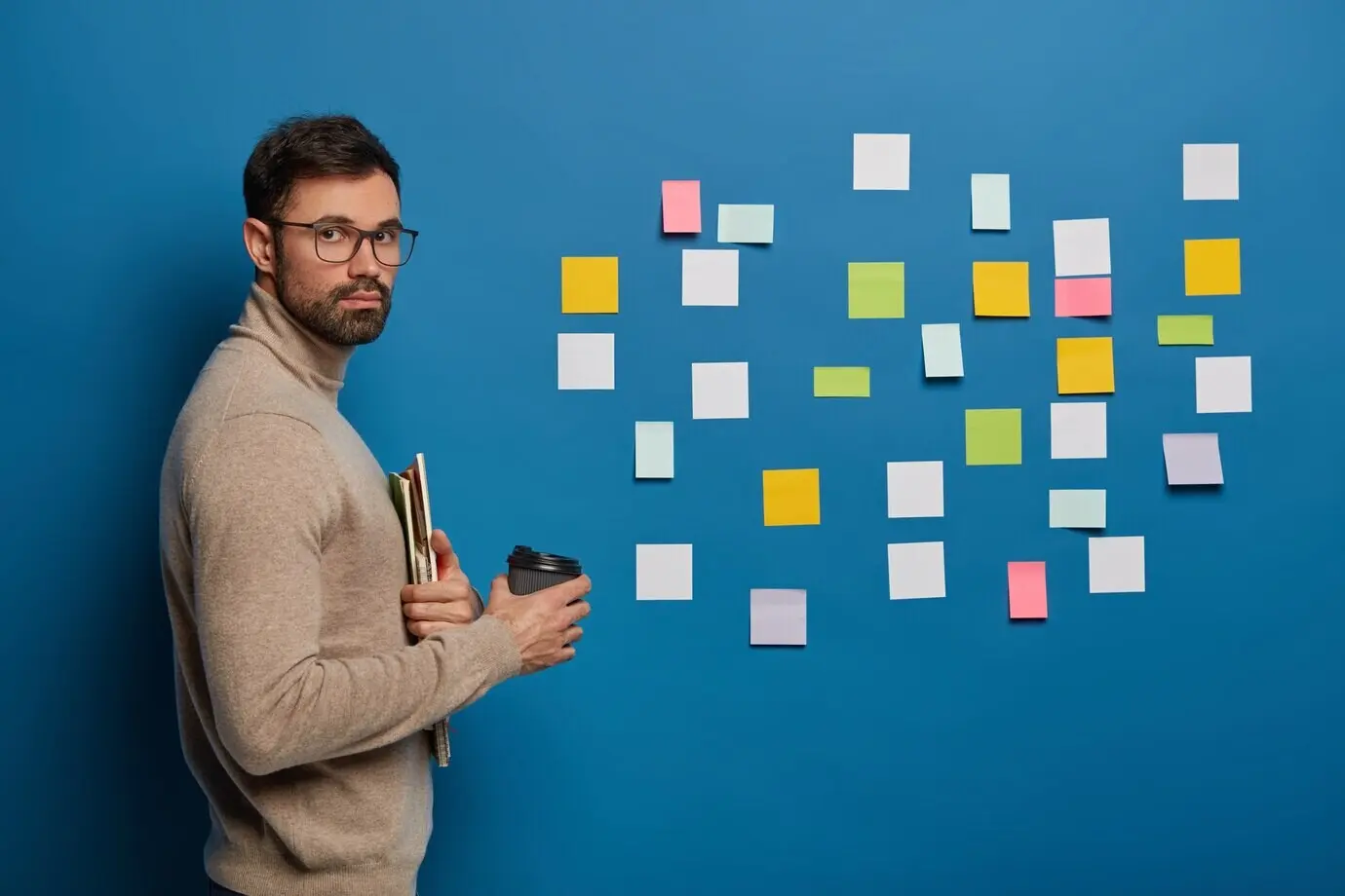 Бородатый студент-мужчина делает перерыв на кофе в помещении, держит одноразовый стакан с горячим напитком и использует стикеры, чтобы запоминать новую информацию.