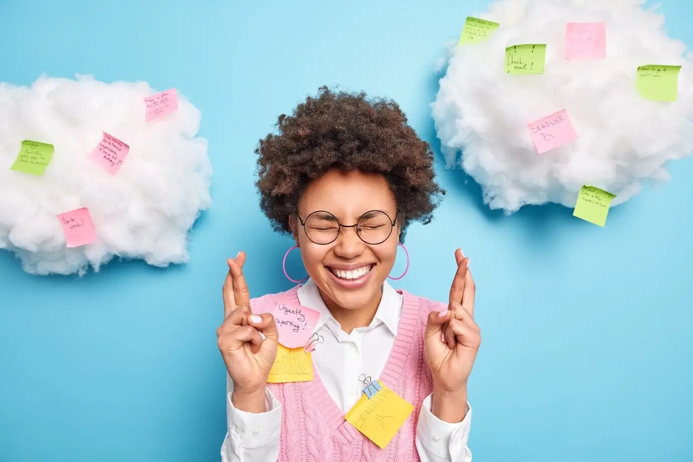 Позитивно настроенный студент с тёмными кудрявыми волосами держит скрещенными пальцы, верит в удачу на экзамене, одет в белую рубашку и жилет, позирует на фоне синей стены.