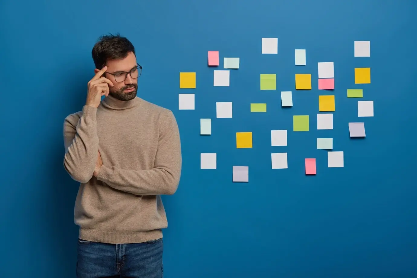 Концепция: люди, работа, размышления. Задумчивый бородатый мужчина держит палец у виска, задумчиво смотрит в сторону и клеит на стену разноцветные стикеры-заметки.
